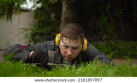 A young man obsessed with work from the special services cuts the lawn with scissors. A man in glasses and headphones lies on the grass and mows the lawn by hand.