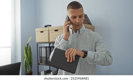 Young man in office checking time on smartwatch while talking on phone, with office furniture and plant in background - Powered by Shutterstock