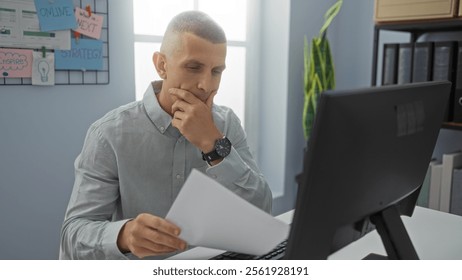 Young man in office working at a computer screen while holding papers with a thoughtful expression.