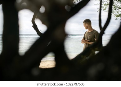 Young man in an olive T-shirt stands near a green tree on the sand beach on the background of the stormy sea and cloudy sky on Gili Trawangan island in Indonesia. View through branches. Horizontal. - Powered by Shutterstock