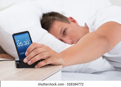 Young Man On Bed Snoozing Alarm Clock On Cell Phone Screen