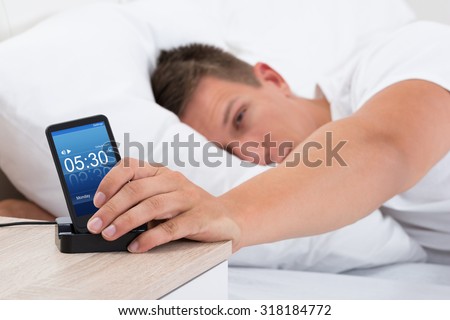 Young Man On Bed Snoozing Alarm Clock On Cell Phone Screen