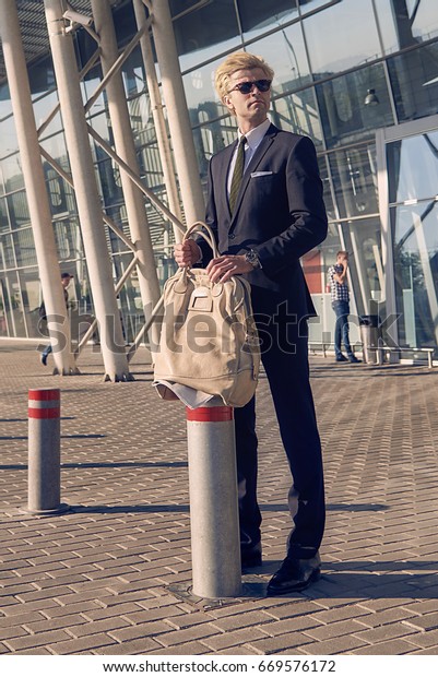 Young man on the\
fashion nice suit with travel bag and newspaper waiting at the\
airport near modern\
building