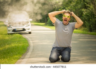 Young man on his knees on the road. He is under stress and pulling his hair out because his car is broken and in smoke. Travel and vacation road problems and assistance concepts. 