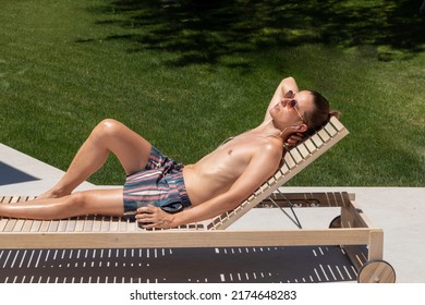 Young Man On Sunglasses And Swimsuit Lying On A Sun Lounger Sun Bathing Close To The Garden Grass