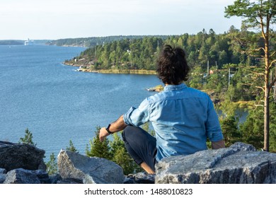 Young man on top rocky shore looking to horizon. Handsome dark haired male have meditation on sea coast. Stressless view of Scandinavia and meditating on blue sky background in peace and zen positive.