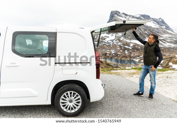 Young man open the trunk of his hired car to\
get some stuff on mountains\
background