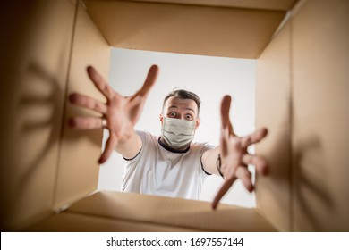 Young Man Opening The Huge Postal Package Wearing Protective Face Mask. Male Model On Top Of Cardboard Box. Food And Goods Contactless Delivery During Coronavirus Quarantine For Isolated People.