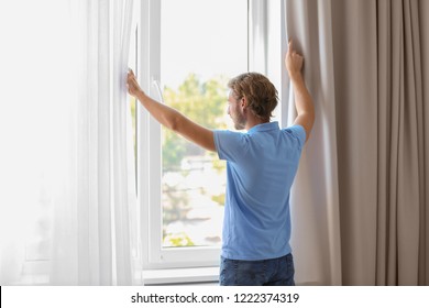Young Man Opening Window Curtains At Home