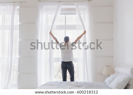 Young man opening window curtains. White color home interior with bed. Rear view
