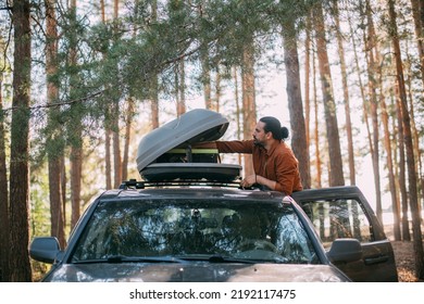 A young man opens the upper tourist trunk of a car in a pine forest on the shore of a lake at sunset. A tourist and an SUV with a trunk on the roof among the trees on a forest road in the sunlight.