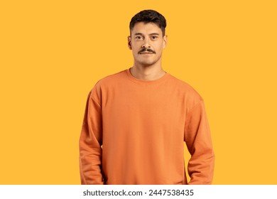 A young man in an orange sweater stands calmly with a neutral facial expression against a solid yellow background