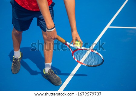 young man in an orange T-shirt and dark blue shorts plays tennis on a blue hard-coated court