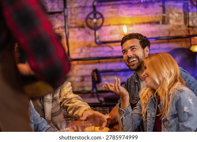 Young man ordering beer while sitting with friends in a stylish industrial pub. Blurred waiter and modern bar interior create authentic nightlife atmosphere.