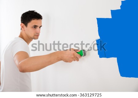 Young man painting wall with blue paint roller at home