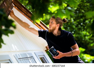 Young Man Painting Wooden House Exterior With Paintbrush. Home Maintenance Concept, Renewing Paint Layer On Outdoors Wooden Wall.