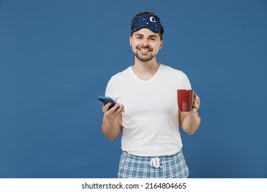 Young man in pajama jam sleep mask rest relax at home hold red cup with milk drink hot decaf coffee tea use mobile cell phone isolated on dark blue background studio Good mood night bedtime concept
