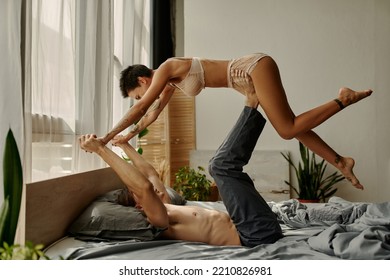 Young Man In Pajama Pants Lifting Tattooed Girlfriend On Bed In Morning