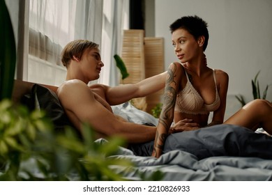 Young Man In Pajama Pants Touching Tattooed Girlfriend In Bra On Bed