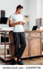 Young Man In Pajamas Holding Cup Of Coffee And Looking At Laptop In Kitchen