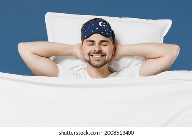 Young man in pajamas jam sleep mask rest relaxing home with lies wrap covered under blanket duvet with closed eyes hands behind neck isolated on dark blue background. Good mood night bedtime concept. - Shutterstock ID 2008513400