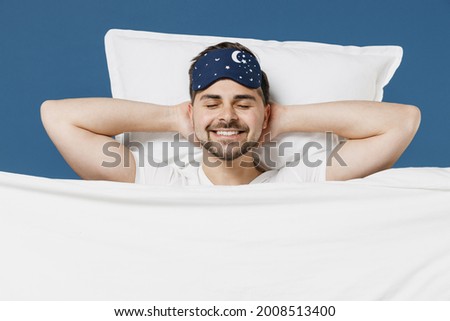 Young man in pajamas jam sleep mask rest relaxing home with lies wrap covered under blanket duvet with closed eyes hands behind neck isolated on dark blue background. Good mood night bedtime concept.