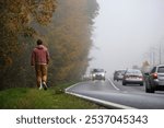 A young man in pants and a jacket walks along the autumn roadside along an autumn forest and an intercity highway along which cars are driving with their headlights on, on a foggy day