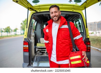 Young man , a paramedic, standing at the rear of an ambulance, by the open doors. He is looking at the camera with a confident expression, smiling, carrying a medical trauma bag on his shoulder.