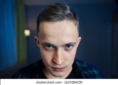 Young Man With Paranoid Schizophrenia Looks Out From Under His Eyebrows With Hatred. An Embittered Guy With Gray Eyes And A Short Haircut Against A Dark Blue Room. 22 Year Old Boy With An Angry Look.