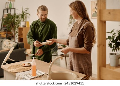 Young man passing dirty plate to his wife standing by kitchen counter for washing after throwing food leftover in trash bin with compost - Powered by Shutterstock