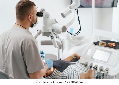 Young Man With Patient Bib On A Dental Chair And A Dentist Who Sits Next To Him. He Looks On His Teeth Using A Dental Microscope And Holds A Dental Bur And A Mirror.