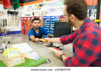 Young Man Paying At The Cashier For His Tools And Paint While Talking With The Hardware Store Worker 