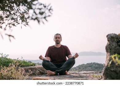 A young man peacefully meditates at sunset in a garden overlooking the mountains and the sea. A Caucasian guy does yoga in simple asanas on a viewpoint in the sunset sunlight - Powered by Shutterstock