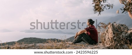A young man peacefully meets the sunset in a garden with a view of the mountains and the sea. A Caucasian guy admires the view of the ocean and hills on a viewpoint in the sunset sunlight