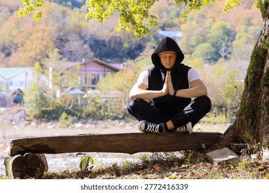 Young man performs prayer mudra sitting on wooden bench by river, black attire against autumn woodland and distant houses.