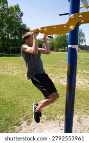 A young man performs pull-ups on an outdoor fitness station. He's wearing athletic attire, and the setting is a grassy field.