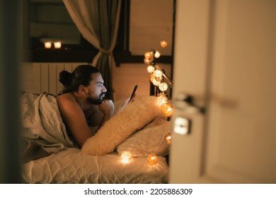 Young man with phone in bed in the evening. A handsome brunette guy lies on the bed before going to bed, looks at the phone. Evening leisure at home - Shutterstock ID 2205886309