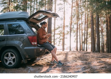 A young man with a phone in his hands next to a car in a pine forest on the shore of a lake at sunset. A tourist with a smartphone and an SUV among the trees on a forest road in the sunlight. - Powered by Shutterstock