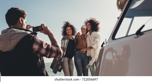 Young Man Photographing Of Female Friends On Seaside. Group Of Young People On Road Trip Taking Pictures.