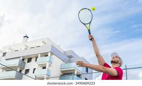 Young man in a pink shirt and white cap serving on an outdoor tennis court with modern buildings in the background. - Powered by Shutterstock