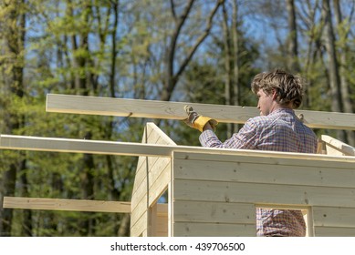 Young Man Placing The Main Wooden Beam To Assemble A Roof Of A Wooden Playhouse Outside In Nature In A DIY Concept.