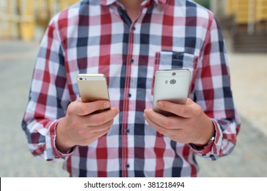 Young Man In A Plaid Shirt With Two Phones In His Hands. Gold And Silver.