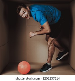 Young Man Playing With Ball In Cardboard Box. Sports And Healthy Lifestyle. Life In Little Cardboard Box. Uncomfortable Life. Personal Spase Concepts. Young Introvert. Man In Sportswear.