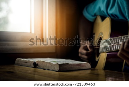 Young man is playing guitar and sing a song from Christian hymn book with bible on wooden table