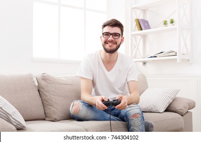 Young Man Playing Video Games At Home. Happy Guy In Glasses Sitting On Couch With Joystick In Hands, Copy Space