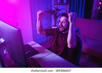 Young man plays computer games at home. The player is celebrating a victory. Neon streamer studio. The guy with the headphones is having fun on the Internet. - Powered by Shutterstock