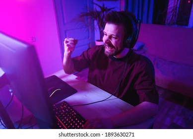 Young Man Plays Computer Games At Home. The Player Is Celebrating A Victory. Neon Streamer Studio. The Guy With The Headphones Is Having Fun On The Internet.