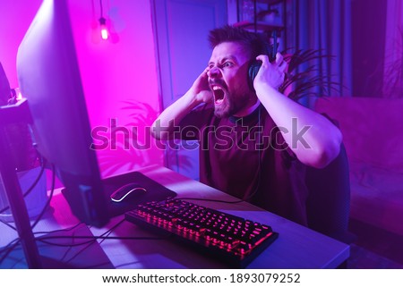 Young man plays computer games at home. The player is upset about the defeat. The streamer is angry during the broadcast. Neon studio setting. Guy surfing the Internet with headphones