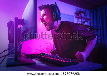 Young man plays computer games at home. The player is upset about the defeat. The streamer is angry during the broadcast. Neon studio setting. Guy surfing the Internet with headphones