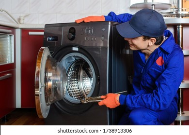 Young Man Plumber Handyman In Blue Overalls Repair/fix The Washing Machine In The Kitchen.